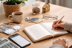 L’image illustre cette approche à travers une scène simple et réaliste : une personne note ses dépenses dans un carnet, entourée d’éléments du quotidien comme un téléphone, une calculatrice, de l’argent liquide et des cartes de crédit.
